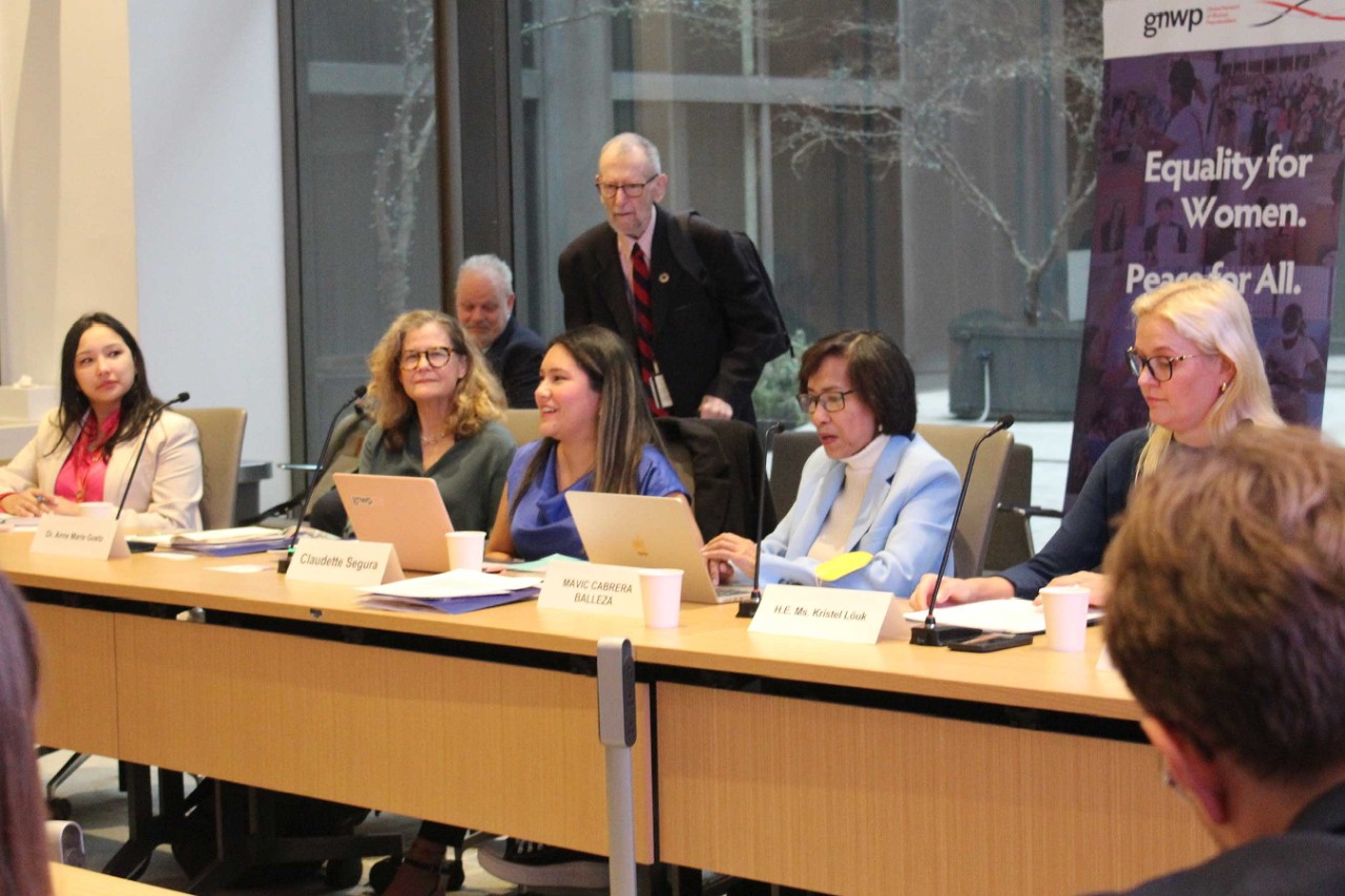 A conference room with people seated at a table, a banner reads "Equality for Women. Peace for All."