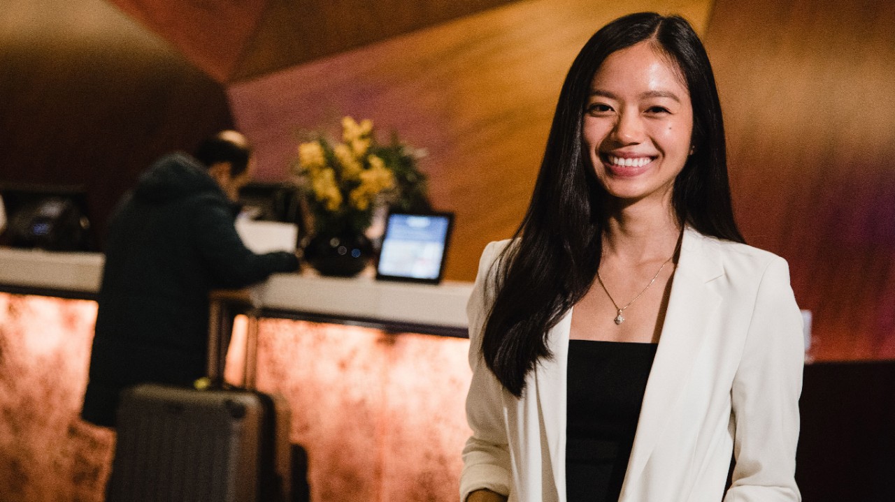 Woman smiles in front of the front desk of a hotel lobby to represent AAS in Hospitality's Your Experience and Our Experience