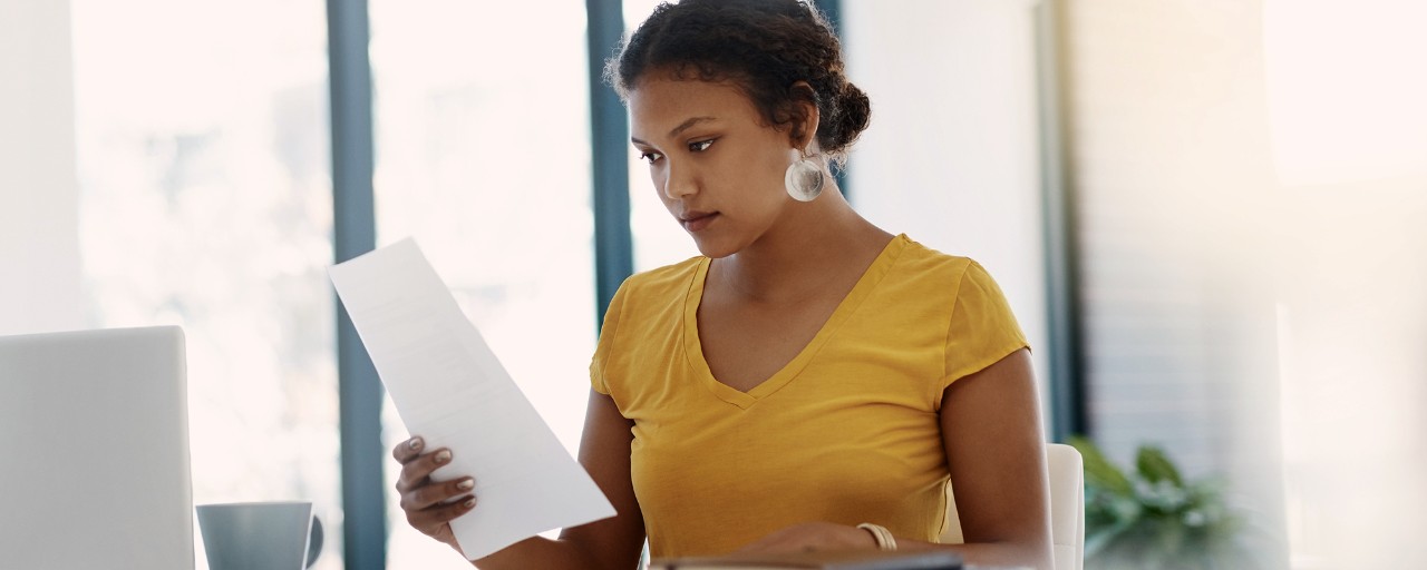 A female online master's degree in translation and interpreting student is sitting behind a computer at a desk while holding and looking at documents.