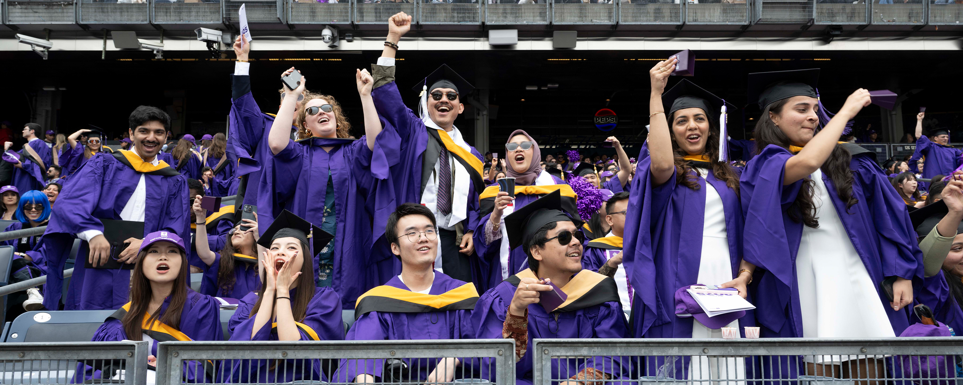 Graduates in purple gowns celebrate at a stadium event, with some cheering and raising their arms.