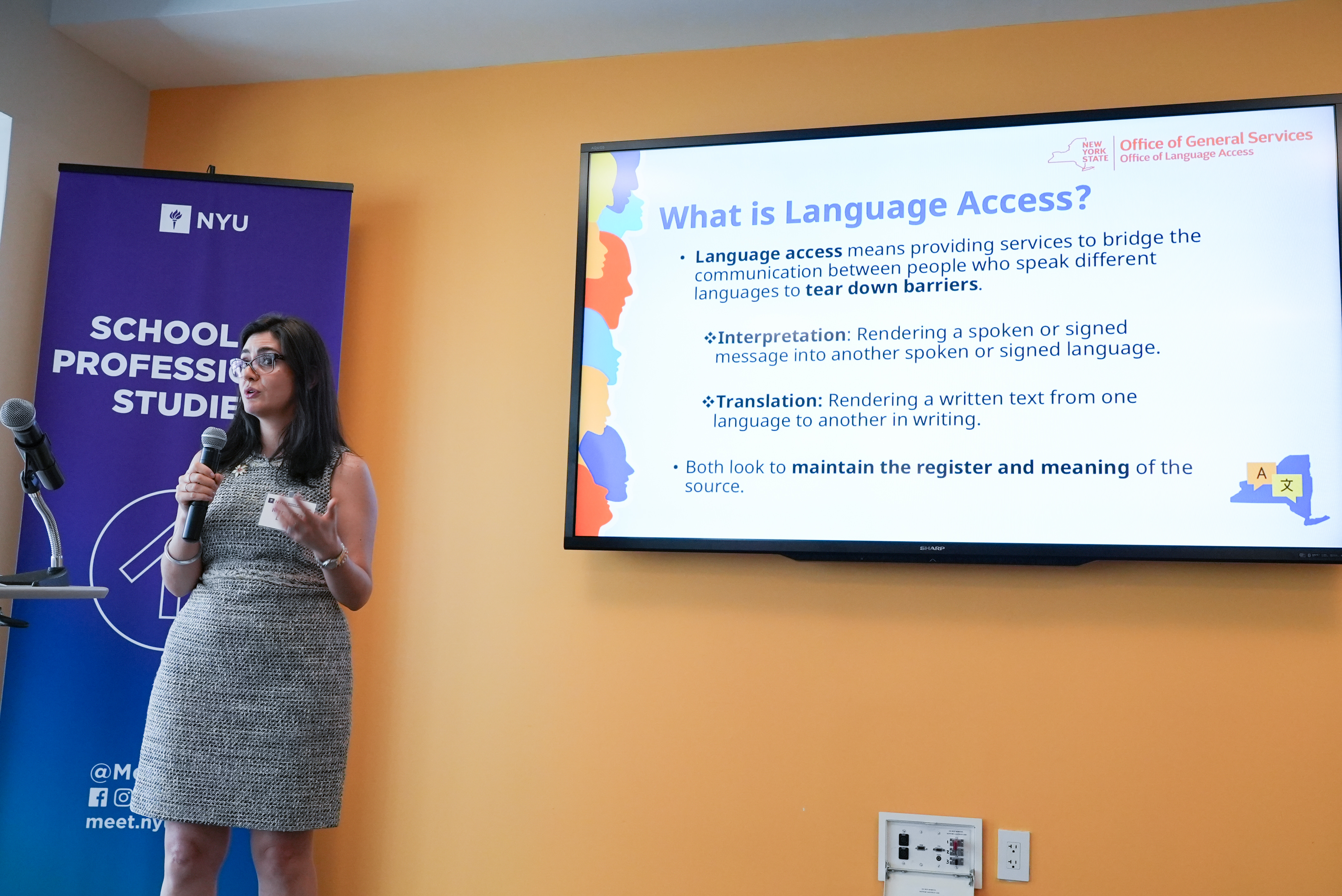 Woman in gray dress holding a microphone in front of TV with a powerpoint on the screen that reads "What is language access?" 